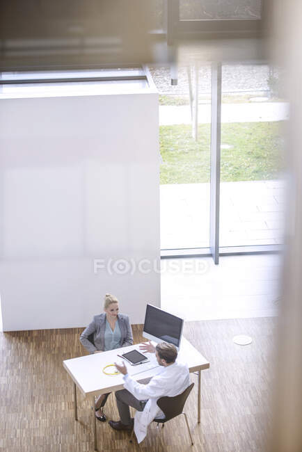Médico masculino y mujer joven sentados en la mesa, discutiendo, vista elevada - foto de stock