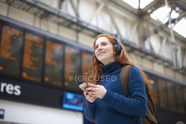 Молодая женщина в наушниках держит смартфон на вокзале — стоковое фото