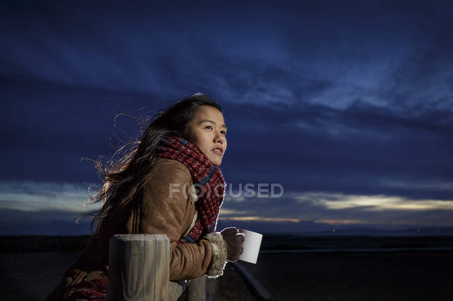 Mujer joven envuelta en una bufanda mirando desde la playa al atardecer - foto de stock
