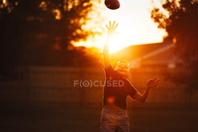 Мальчик практикует американский футбол в саду и тянется ловить мяч — стоковое фото