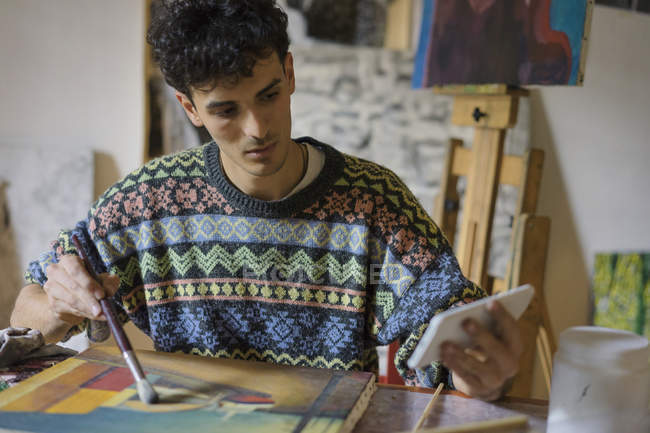 Männlicher Künstler blickt auf Smartphone, während er im Atelier Leinwand malt — Stockfoto