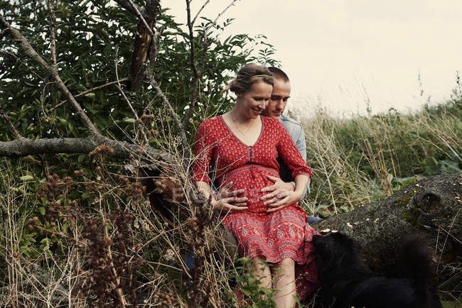 Романтическая беременная пара сидит на стволе дерева — стоковое фото