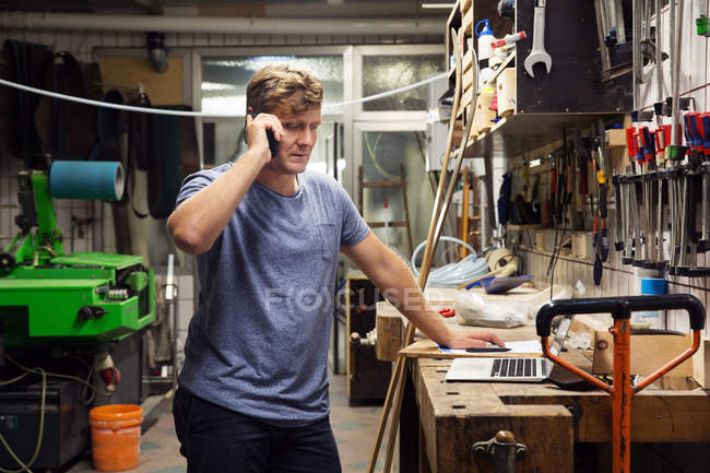Mann in Werkstatt benutzt Smartphone und schaut auf Laptop — Stockfoto