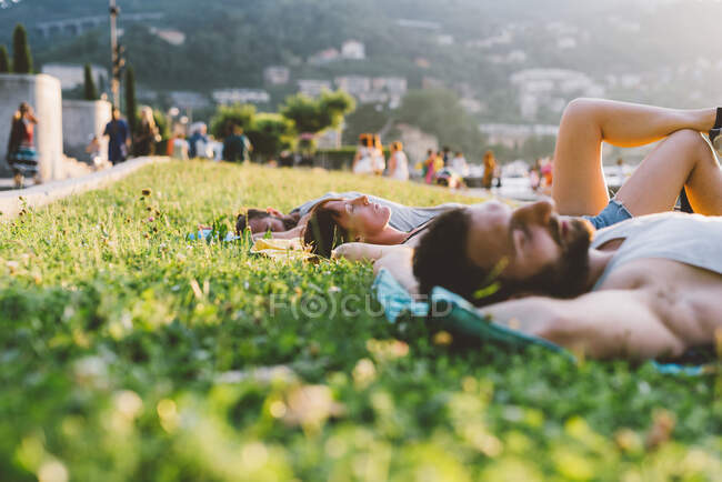 Parejas jóvenes tumbadas en la hierba frente al mar, Lago de Como, Lombardía, Italia - foto de stock