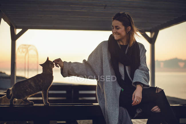 Жінка погладжує кота на лавці на заході сонця — стокове фото