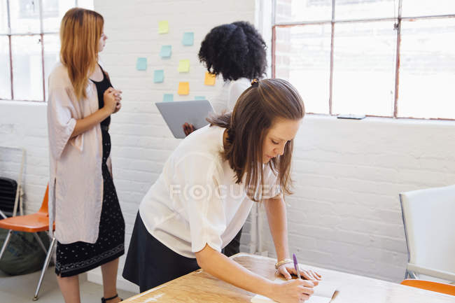 Женщина в офисе пишет в блокноте и два коллеги на заднем плане — стоковое фото