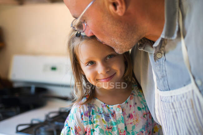 Portrait of girl being kissed on forehead by father in kitchen — Stock Photo