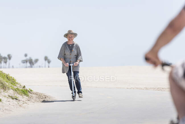 Senior donna guida spingere scooter sulla spiaggia — Foto stock
