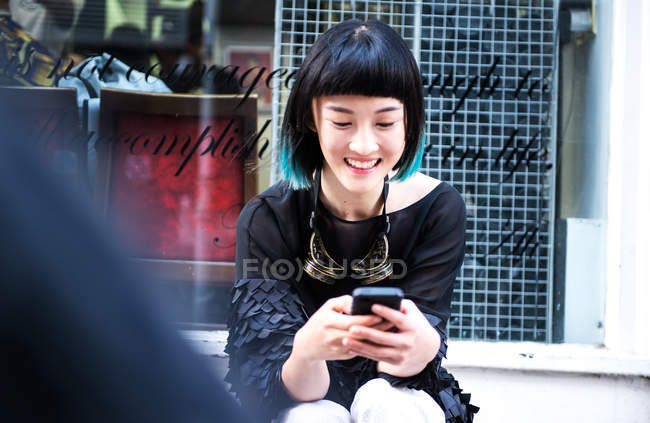 Junge stylische Frau sitzt vor dem Geschäft und schaut auf ihr Smartphone — Stockfoto