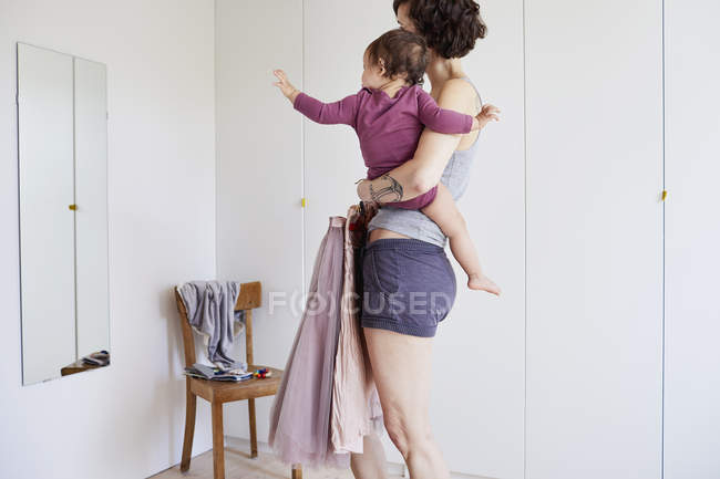 Mãe segurando bebê menina e roupas contra espelho — Fotografia de Stock