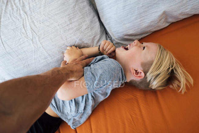 Boy lying on bed being tickled by father's hand — Stock Photo