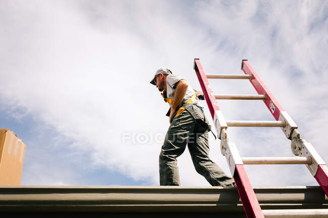 Trabajador en el techo de la casa, escalera inclinada hacia arriba contra el lado de la casa, vista de ángulo bajo - foto de stock