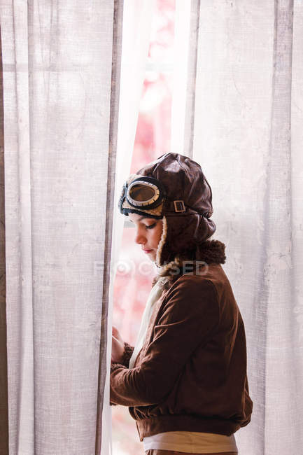 Girl looking out through window curtain wearing pilot costume for Halloween — Stock Photo