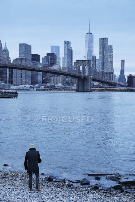 Hombre mirando sobre Brooklyn Bridge y Lower Manhattan Skyline desde riverbank, Nueva York, EE.UU. - foto de stock