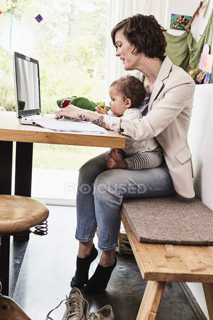 Мать держит на руках маленькую дочь и работает на ноутбуке — стоковое фото