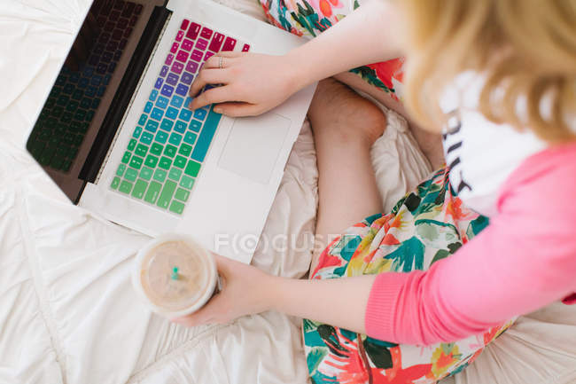 Jeune femme assise sur le lit avec tasse de café à emporter et ordinateur portable — Photo de stock