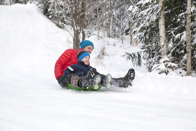 Man and son tobogganing down hill in snow covered forest — Stock Photo