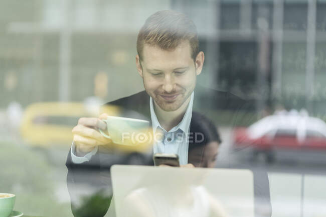 Empresario sentado en la cafetería, bebiendo café, usando teléfono inteligente, visto a través de la ventana - foto de stock