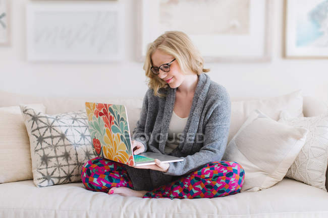 Mujer joven sentada en el sofá y escribiendo en el portátil - foto de stock