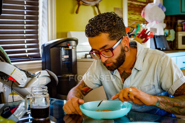 Молодой человек в очках ест за кухонным столом — стоковое фото