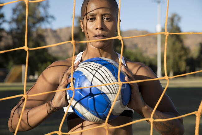 Retrato de mujer detrás de la red de gol de fútbol sosteniendo el fútbol mirando a la cámara. - foto de stock