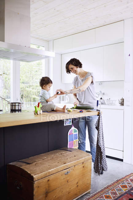 Baby-Mädchen sitzt auf Küchentisch, während Mutter Gurke schneidet — Stockfoto