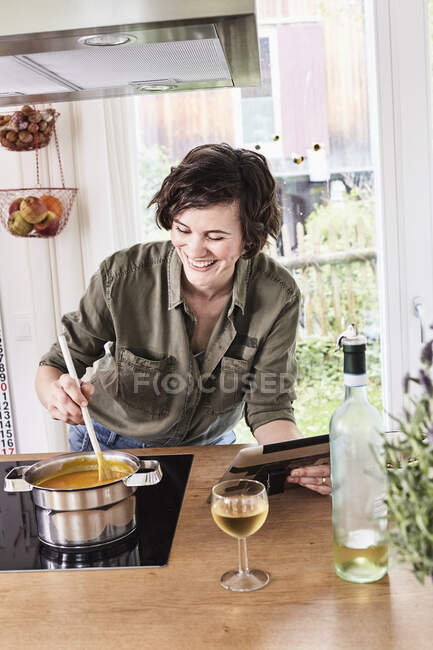 Mid adult woman stirring pot on stove in kitchen, holding digital tablet, laughing — Stock Photo