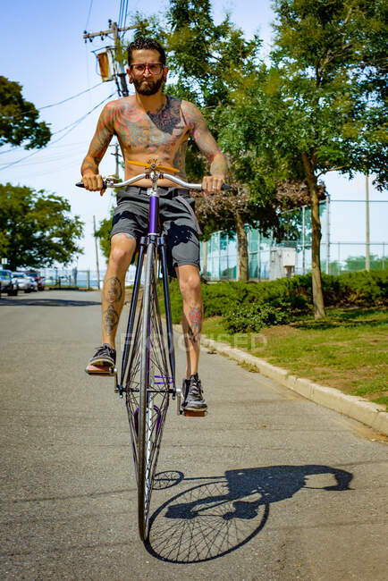 Retrato de joven tatuado ciclismo carretera suburbana en penny farthing - foto de stock