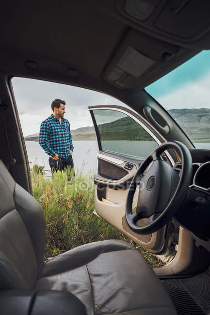 Mid adult man standing beside Dillon Reservoir, view through parked car, Silverthorne, Colorado, USA — Stock Photo