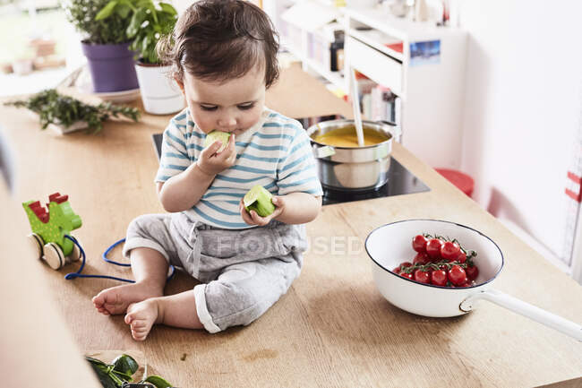 Baby girl sitting on kitchen counter, eating cucumber — Stock Photo