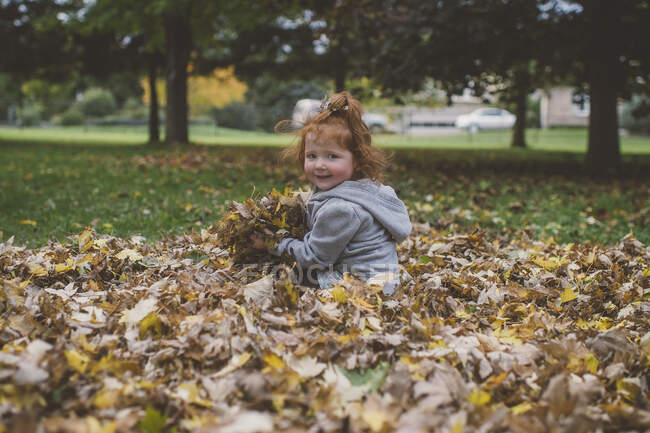 Portrait of red haired female toddler in park with bundles of autumn leaves — Stock Photo