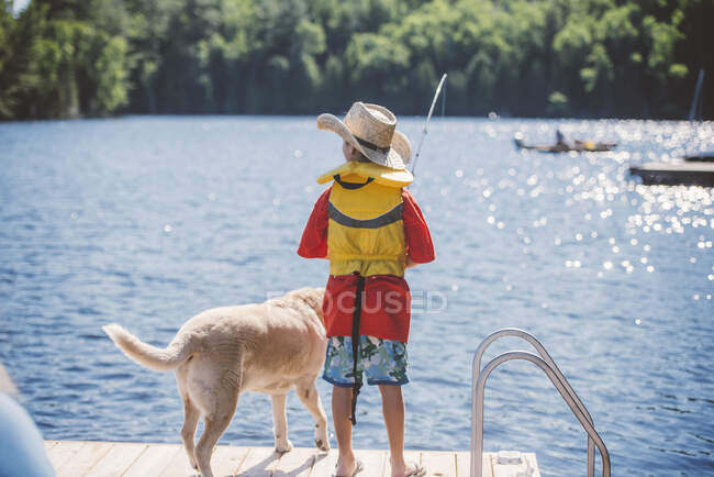 Rear view of dog and boy in cowboy hat fishing from lake pier — Stock Photo