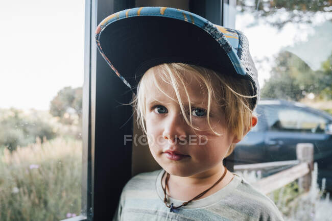 Retrato de niño rubio usando gorra de béisbol por ventana - foto de stock