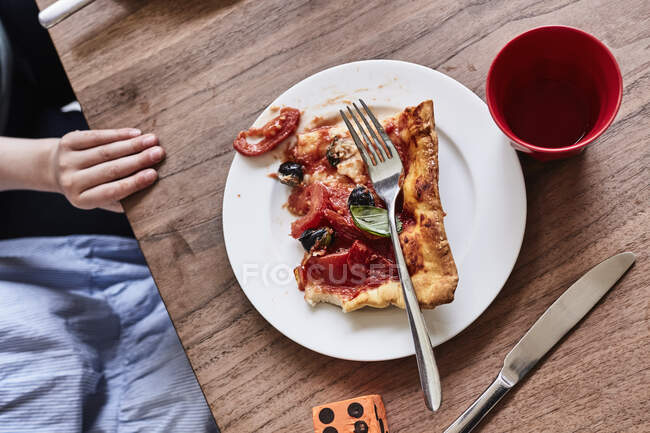 Slice of pizza on white plate, child's hand on table, mid section, elevated view — Stock Photo