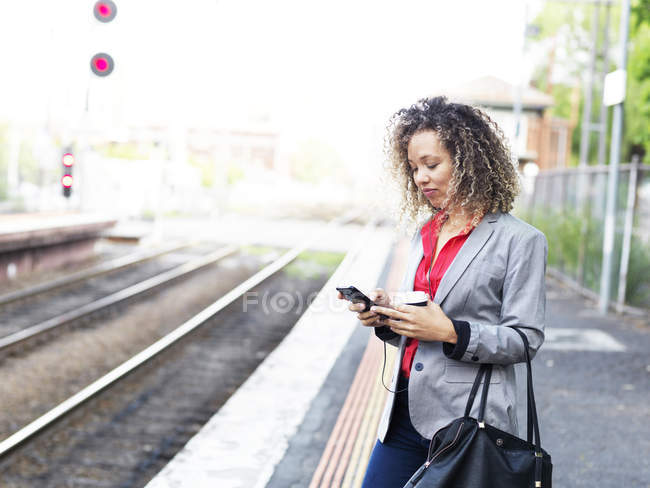 Femme adulte moyenne debout sur la plate-forme du train, en utilisant un smartphone, tenant une tasse de café jetable — Photo de stock