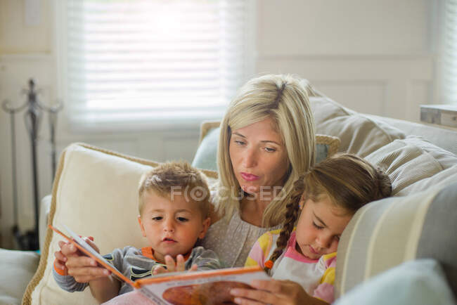 Femme adulte moyenne lisant avec sa fille et son fils en bas âge sur le canapé — Photo de stock