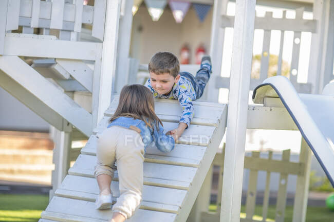 Menina e menino na pré-escola, ajudando a mão a rastejar rampa na moldura de escalada no jardim — Fotografia de Stock