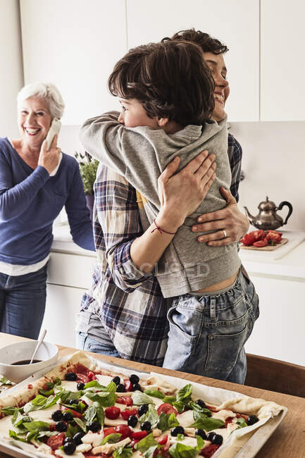 Mutter und Sohn umarmen sich in der Küche, Großmutter im Hintergrund mit Smartphone — Stockfoto