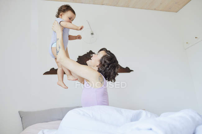 Mãe sentada na cama e segurando bebê menina no ar — Fotografia de Stock