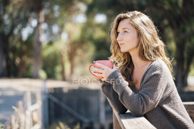 Молода жінка спирається на паркан і тримає чашку кави — стокове фото