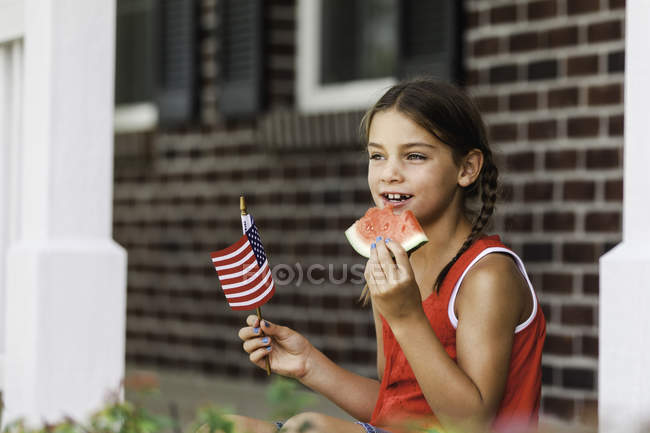 Menina jovem ao ar livre, segurando pequena bandeira americana, comendo fatia de melancia — Fotografia de Stock
