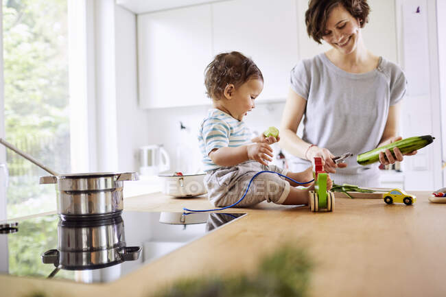 Baby girl sitting on kitchen counter while mother slicing cucumber — Stock Photo