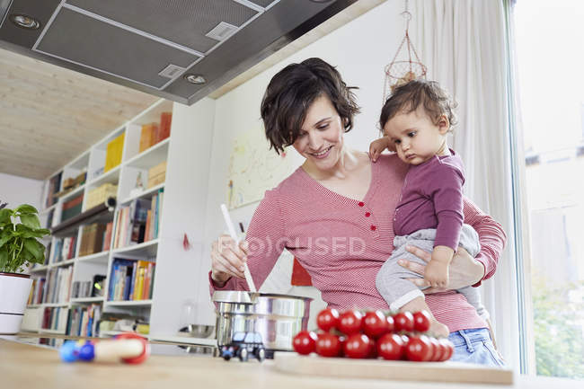 Mother holding baby girl in kitchen while cooking — Stock Photo