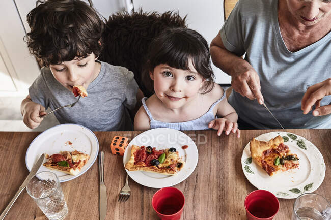 Grand-mère assise à la table de la cuisine avec petits-enfants, manger de la pizza, vue surélevée — Photo de stock