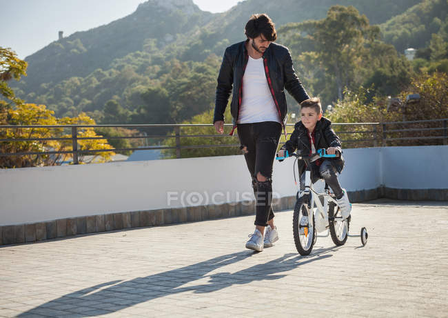 Young boy riding bike with stabilizers while father walking beside him — Stock Photo