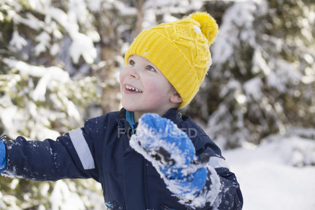 Menino de chapéu de malha amarelo olhando para cima na floresta nevada — Fotografia de Stock