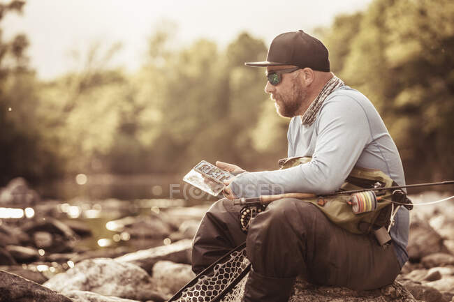 Pescador sentado en las rocas del río mirando el teléfono inteligente, Mozirje, Brezovica, Eslovenia - foto de stock