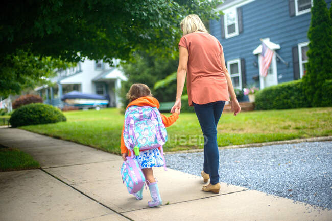 Mujer adulta caminando con su hija en la acera suburbana, vista trasera - foto de stock