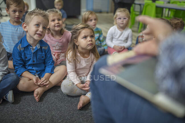 Young children sitting on carpet in classroom, listening to teacher at front of class — Stock Photo