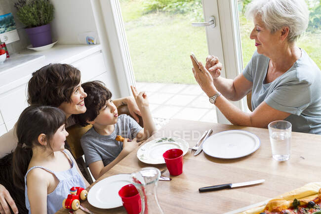 Бабушка сидит на кухне, фотографирует взрослую дочь и внуков, используя смартфон — стоковое фото